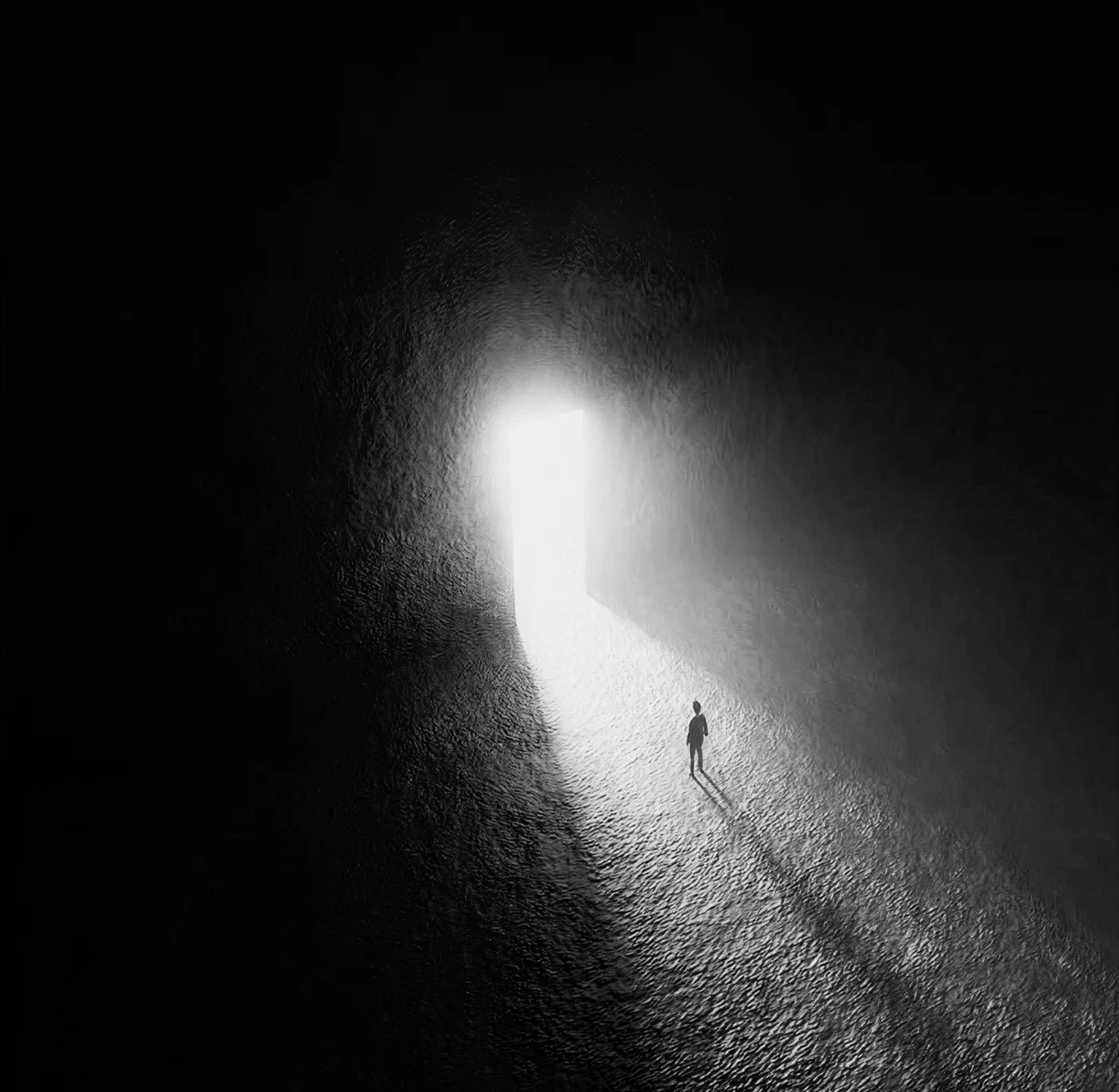 Black-and-white overhead view of a lone figure walking out of a lit cave mouth onto a bright path.