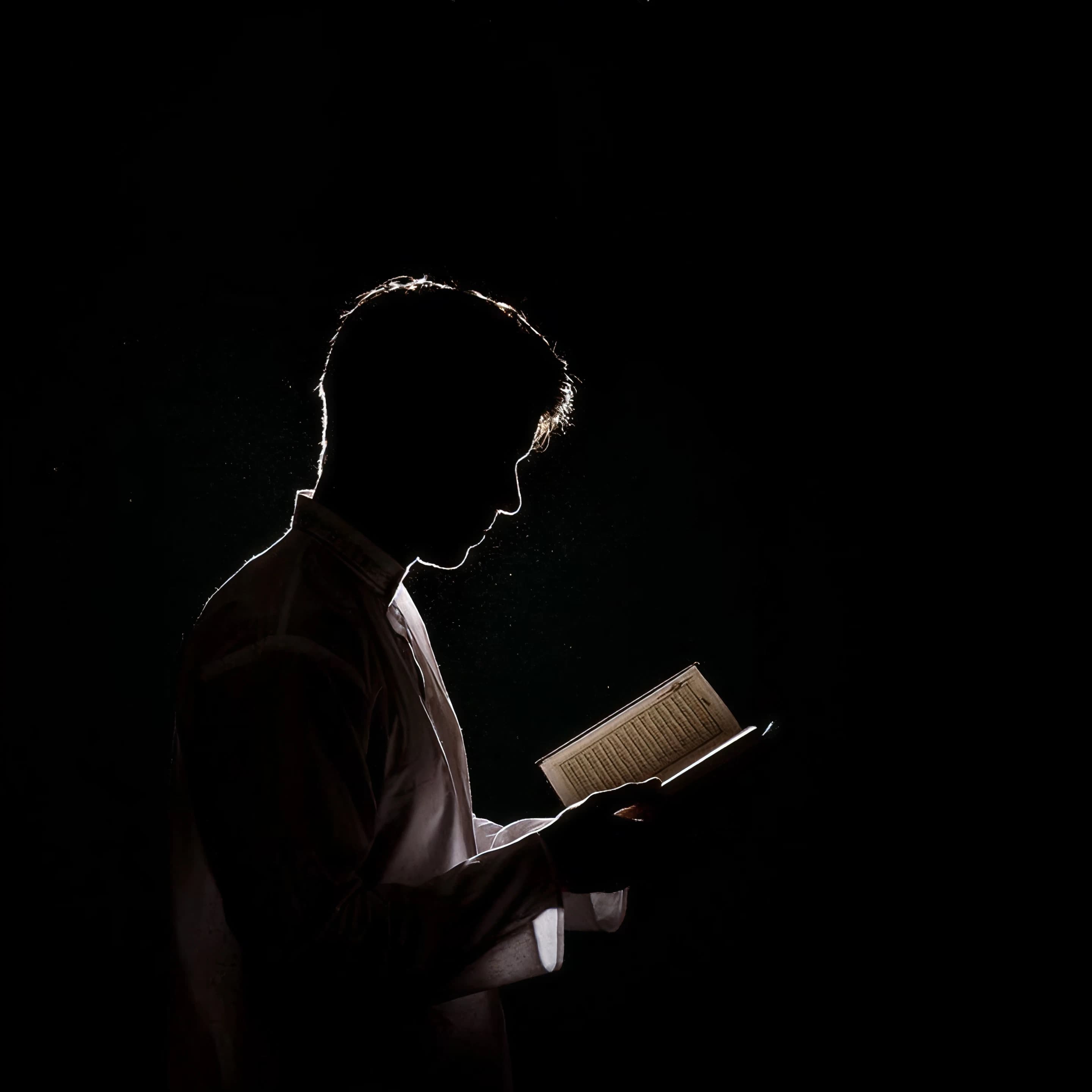 A man in white robes reads from an open Qur'an, rim-lit against a black background.