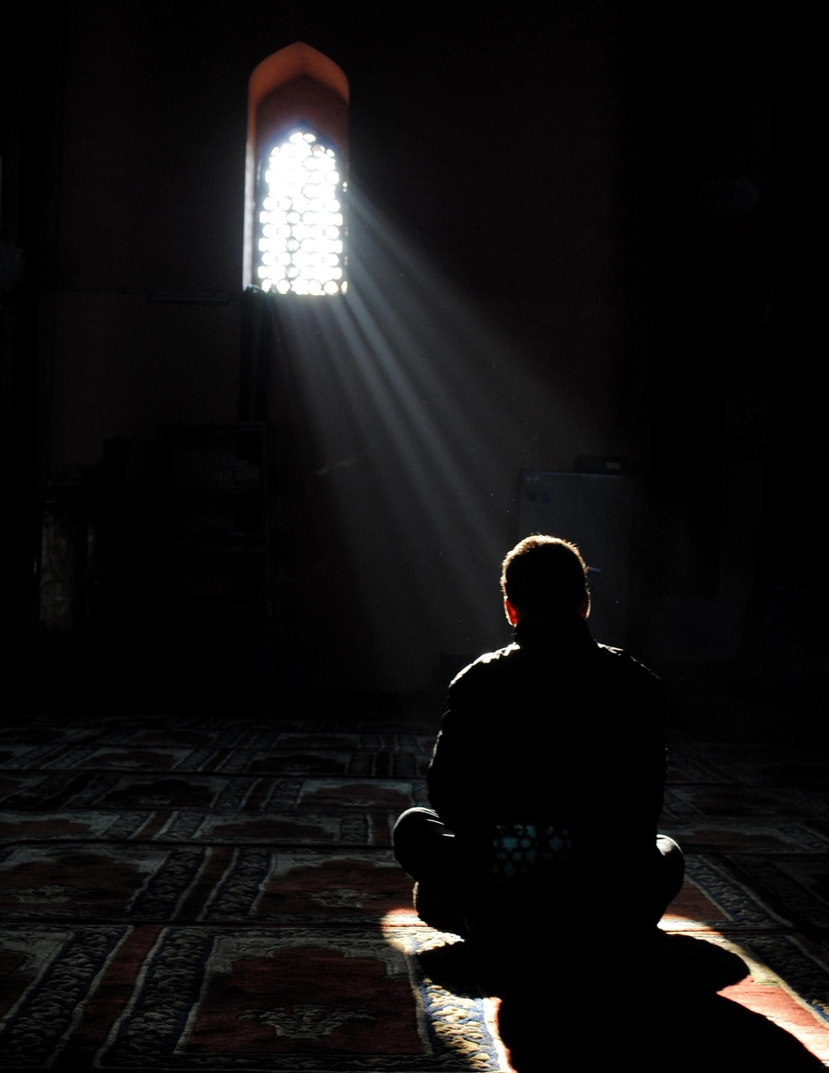 A man sits with his back to camera on a red patterned prayer carpet in a near-black mosque interior, in silhouette under hard vertical rays from one small pierced-screen window.