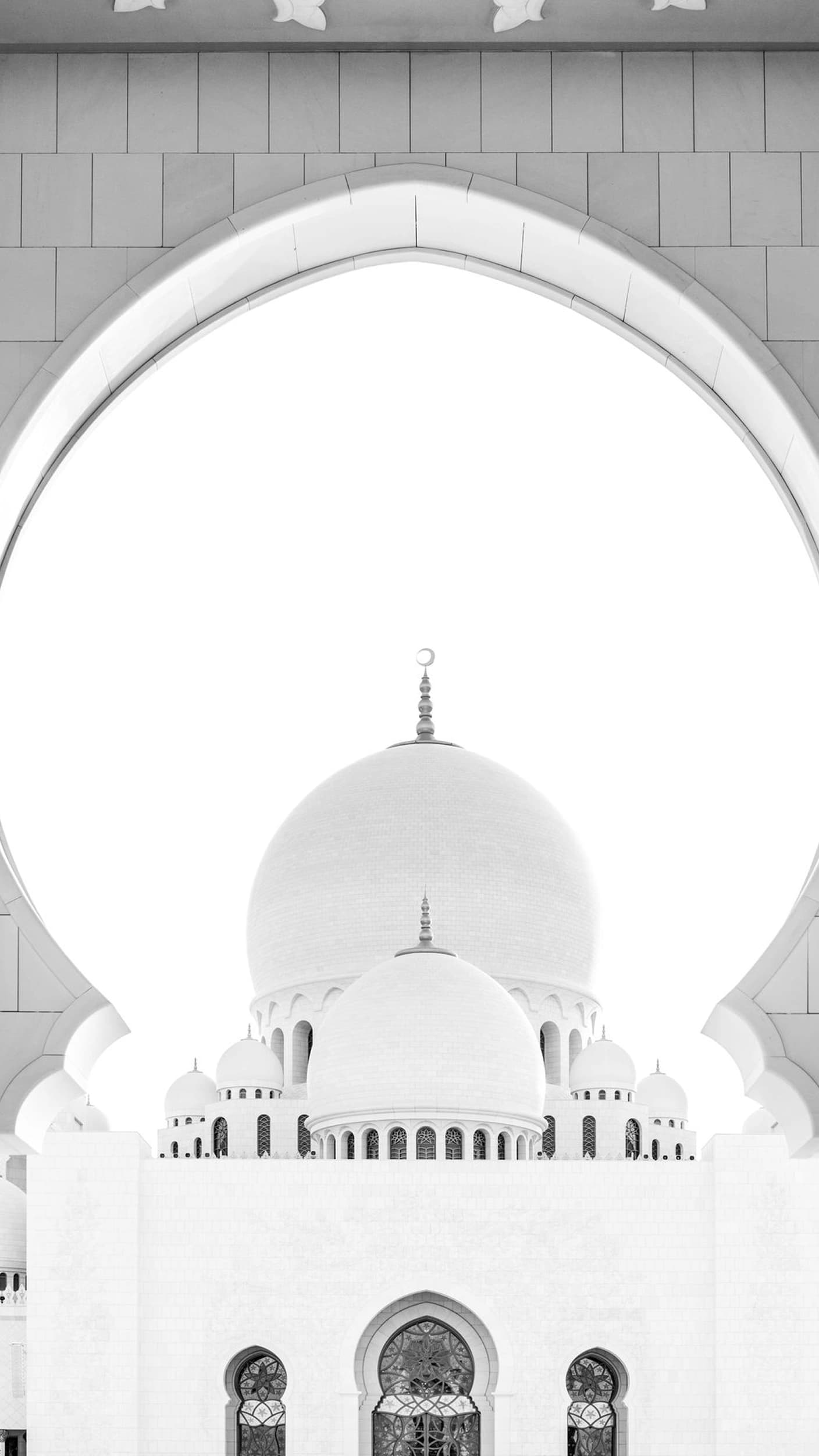 A white mosque dome with a crescent finial, flanked by smaller domes and an arcaded gallery, seen framed through a white pointed arch.