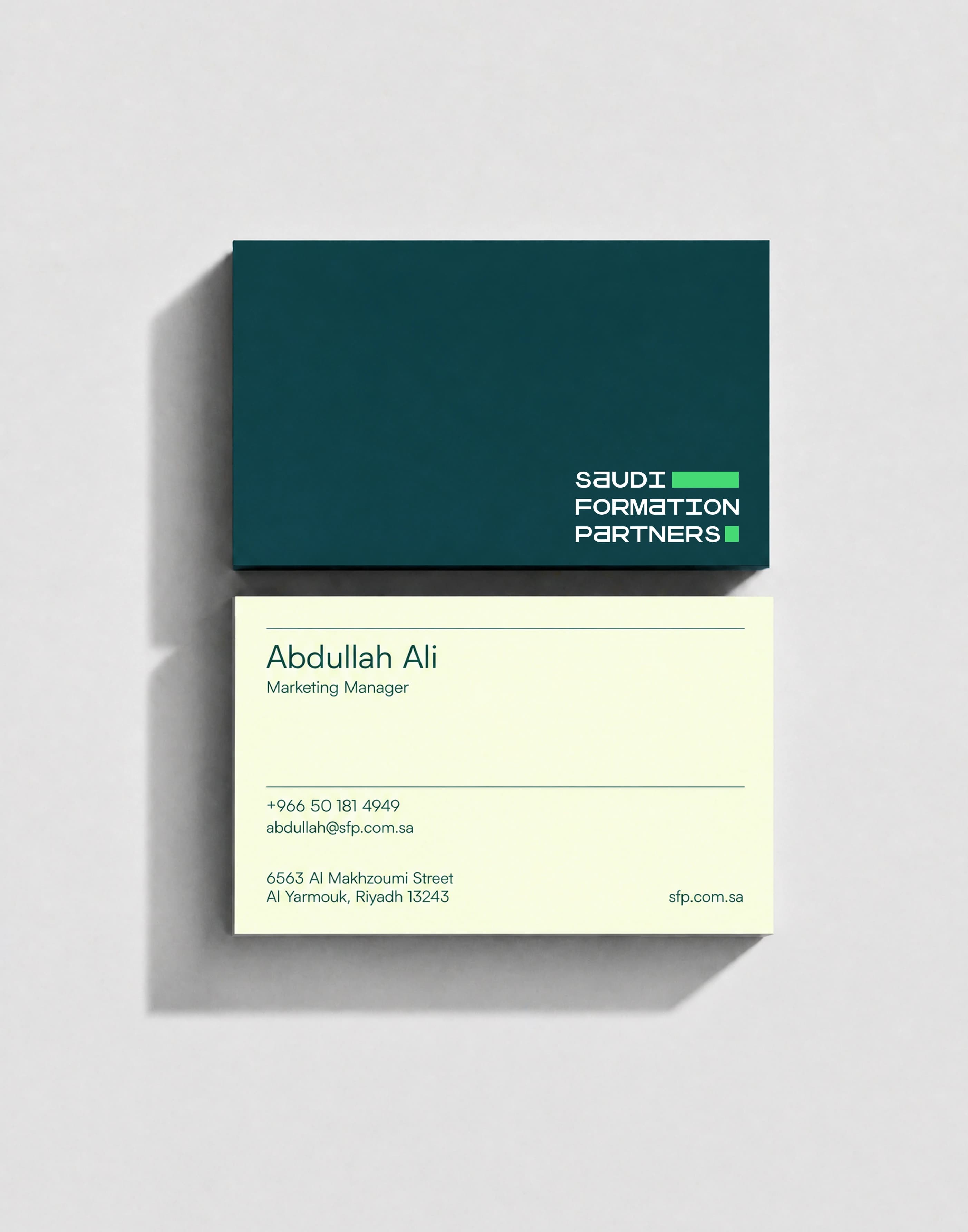 A pair of business cards photographed flat from above on a light grey surface, the dark teal back carrying the wordmark resting on the cream front, which shows a name, job title, phone number, email address and a Riyadh street address.