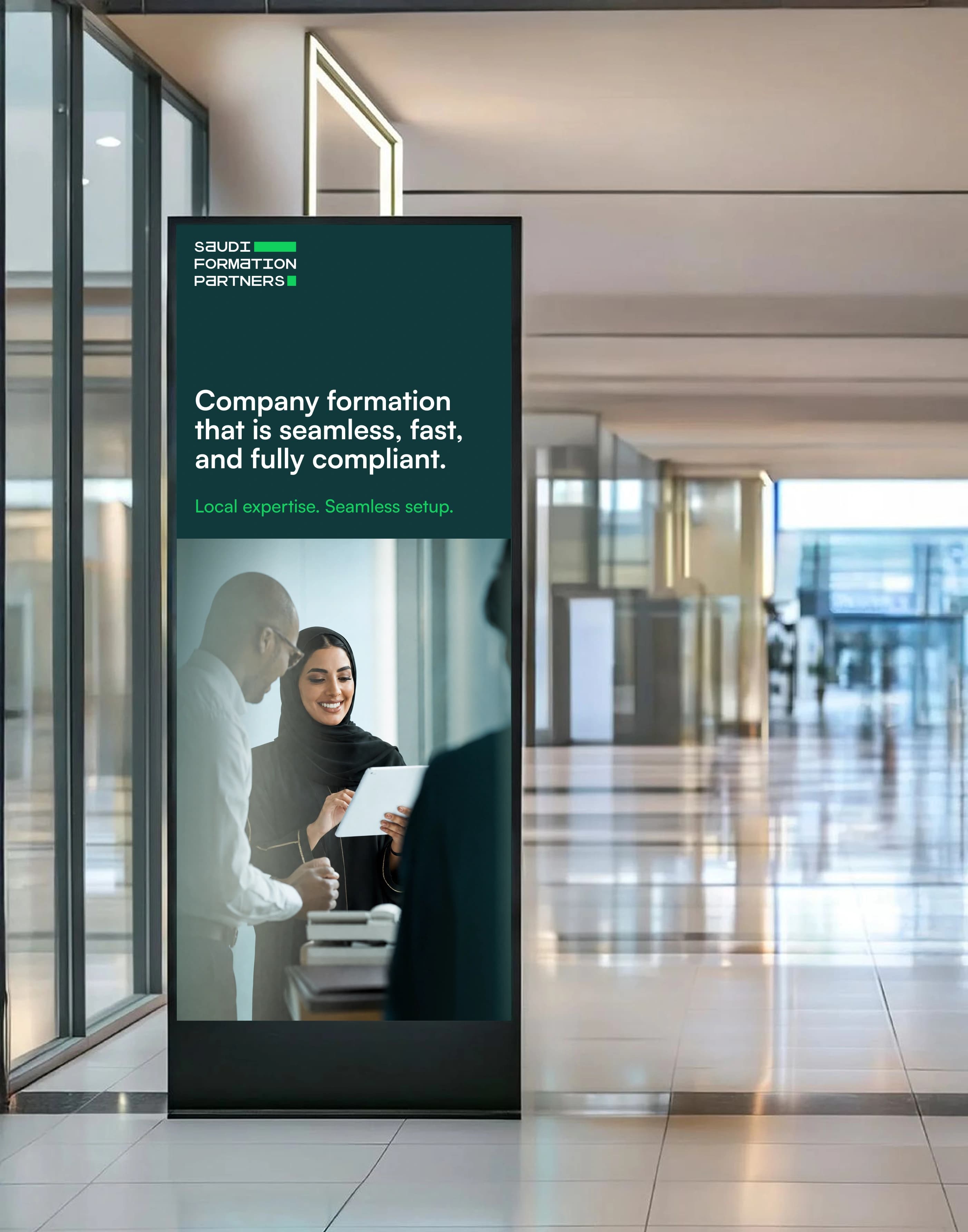 A portrait digital advertising kiosk standing in a bright glass-walled lobby corridor, its dark green screen carrying the Saudi Formation Partners wordmark and a headline about seamless company formation above a photograph of two colleagues, one holding a tablet.