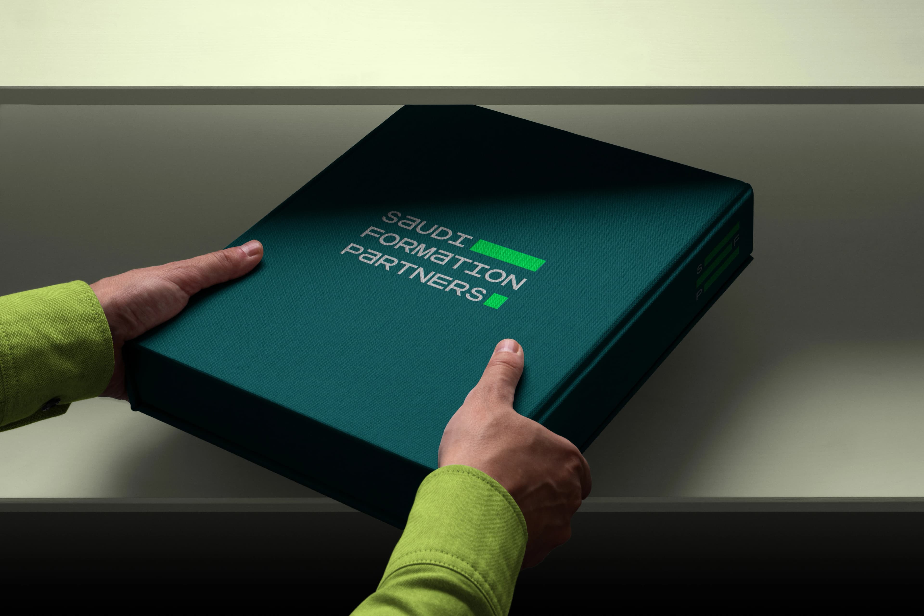 A hand in a lime-green sleeve lifts the lid from a dark teal cloth presentation box printed with the Saudi Formation Partners wordmark.