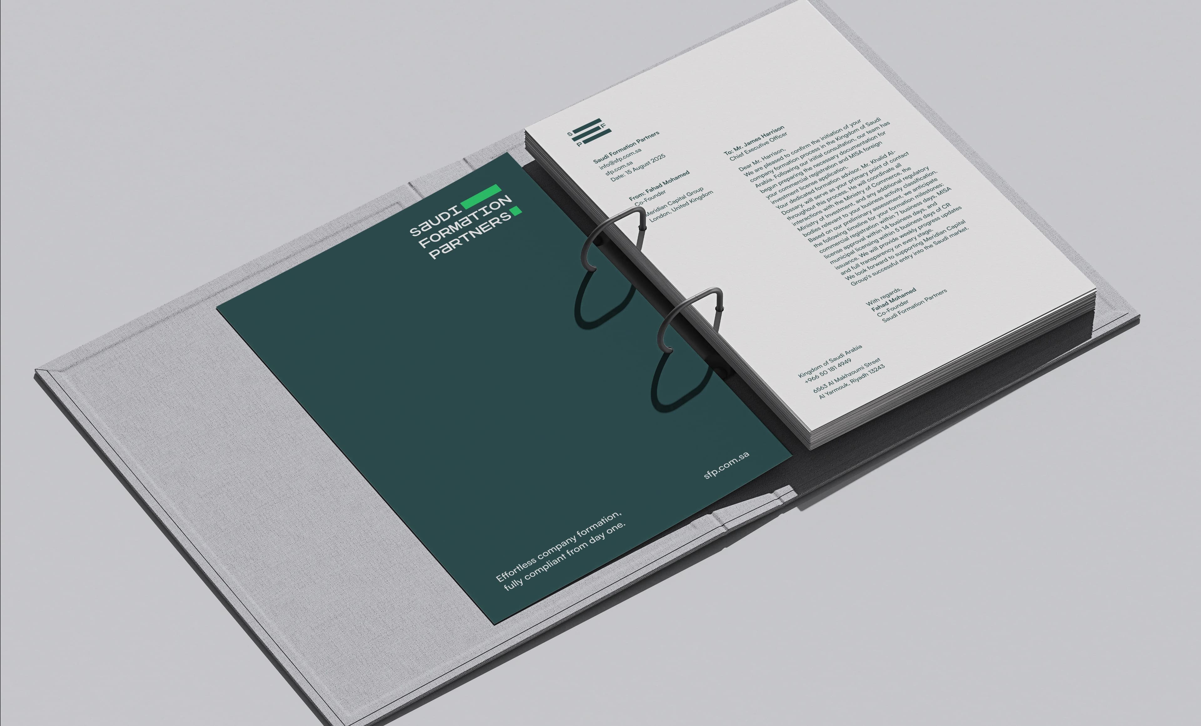 An open two-ring linen binder lying flat on a grey surface, with a punched stack of cream Saudi Formation Partners letterhead pages on its right leaf and a loose dark teal card printed with the company tagline resting on the inside front cover at the left.