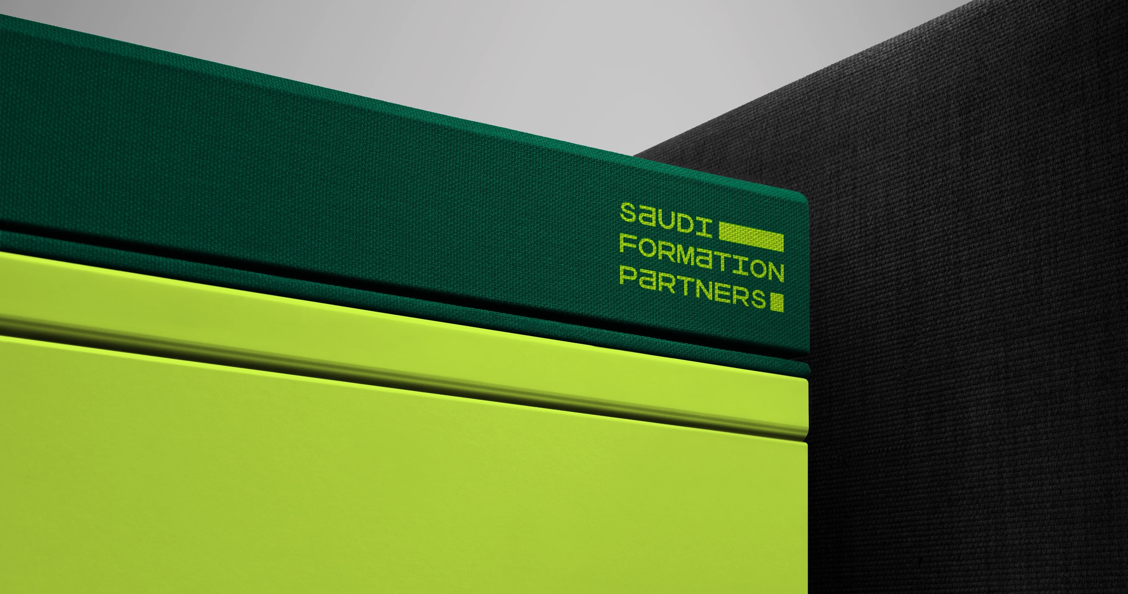 A close crop of the corner of a stack of rigid boxes: a dark green canvas-wrapped box printed with the lime Saudi Formation Partners wordmark rests above two smooth lime-green boxes, against a black woven backdrop.