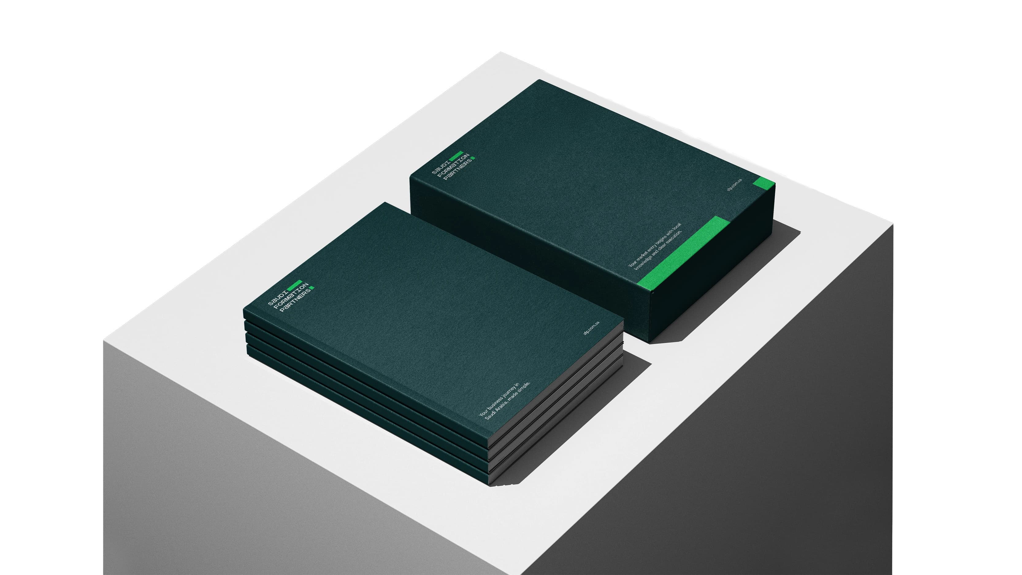 An angled studio shot of a stack of dark teal softcover booklets, their page edges showing, beside a rigid dark teal presentation box with bright lime side bands carrying one more booklet on its lid, on a white plinth against a grey backdrop.