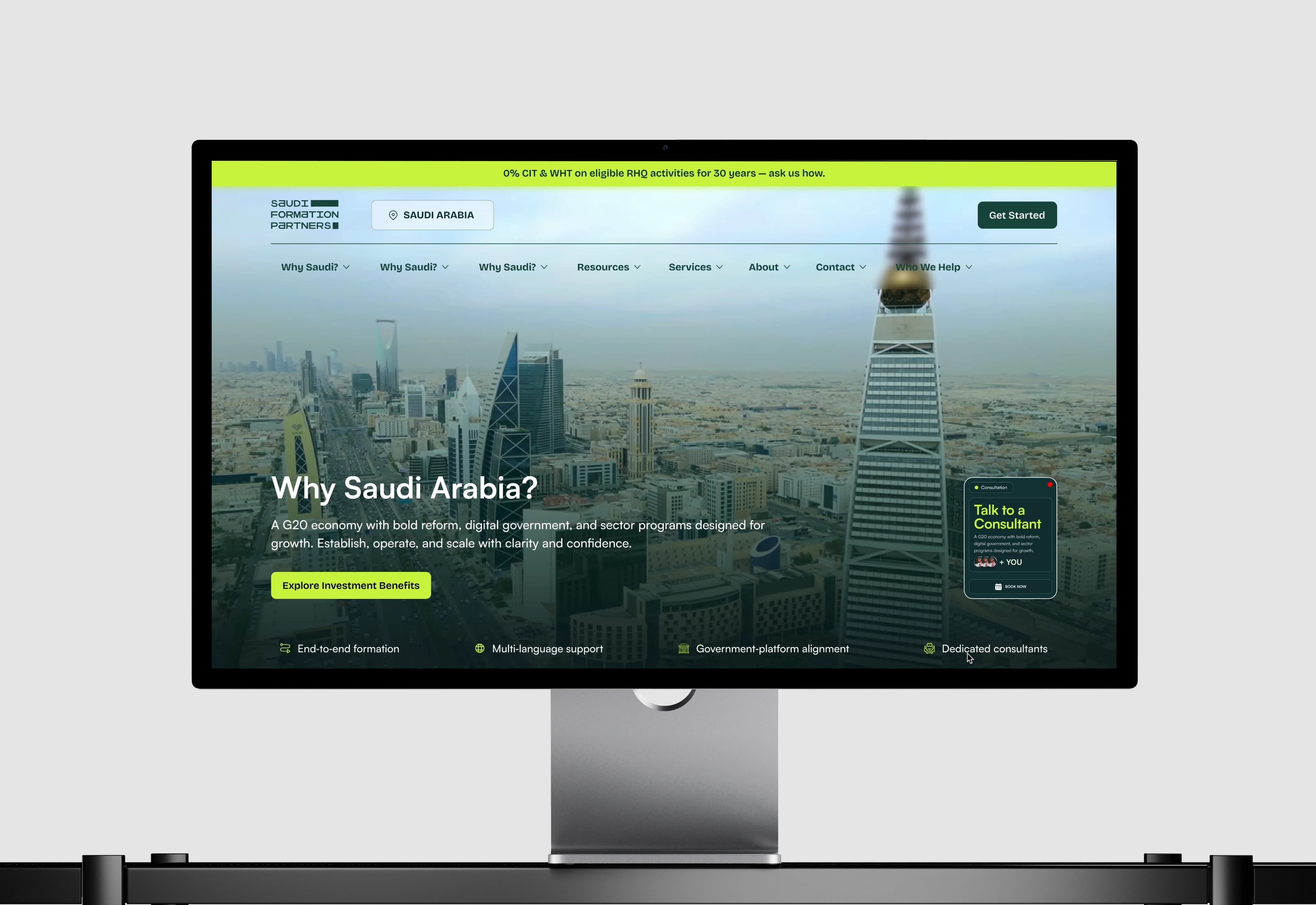 The Saudi Formation Partners website displayed on a studio desktop monitor, a lime announcement bar above the header and an aerial Riyadh skyline hero headed Why Saudi Arabia, with a lime Explore Investment Benefits button and a floating Talk to a Consultant card at the right.