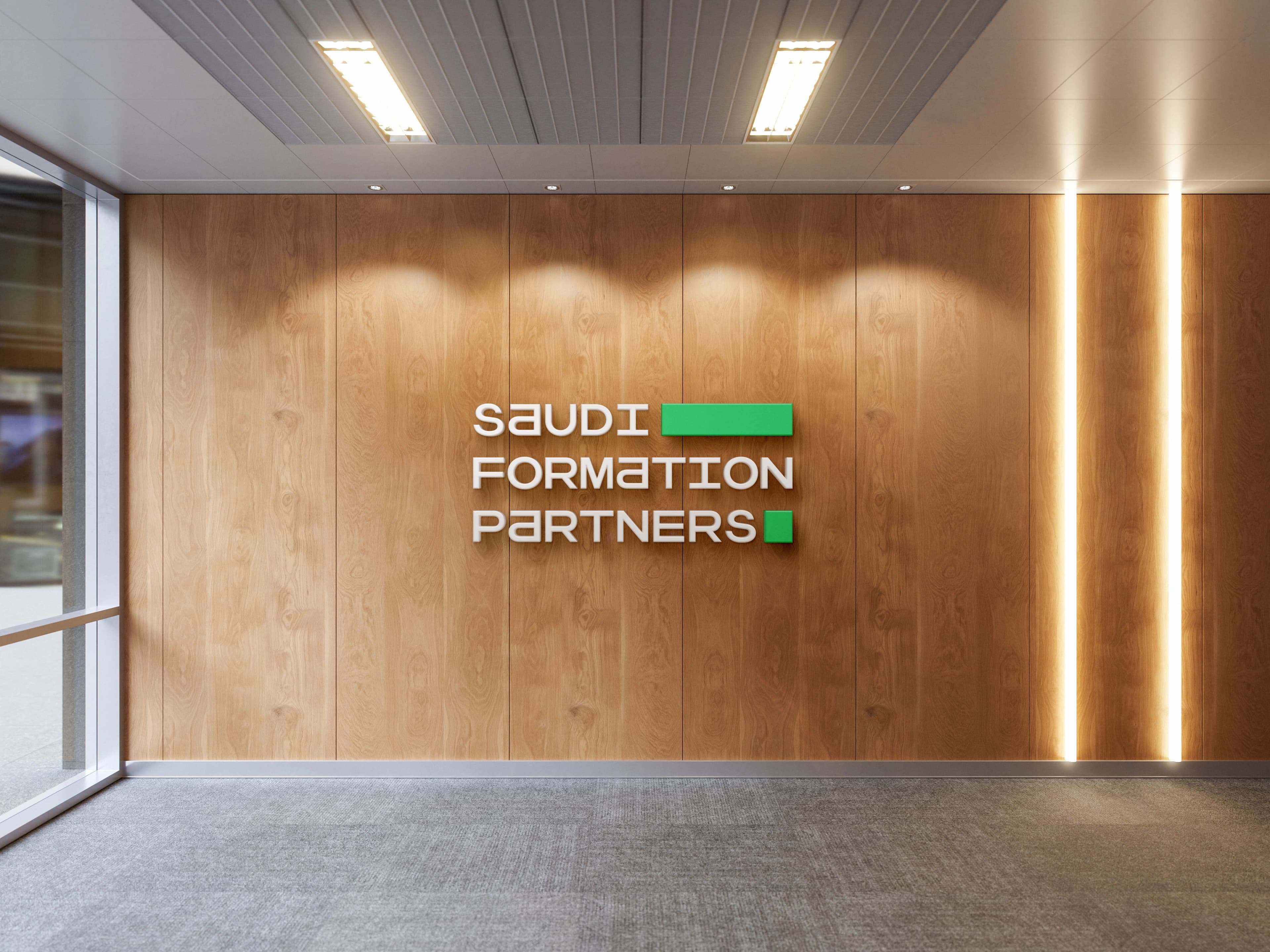 An office reception interior where dimensional white Saudi Formation Partners lettering is mounted on a slatted wood wall, lit by downlights, with glazing along the left edge.