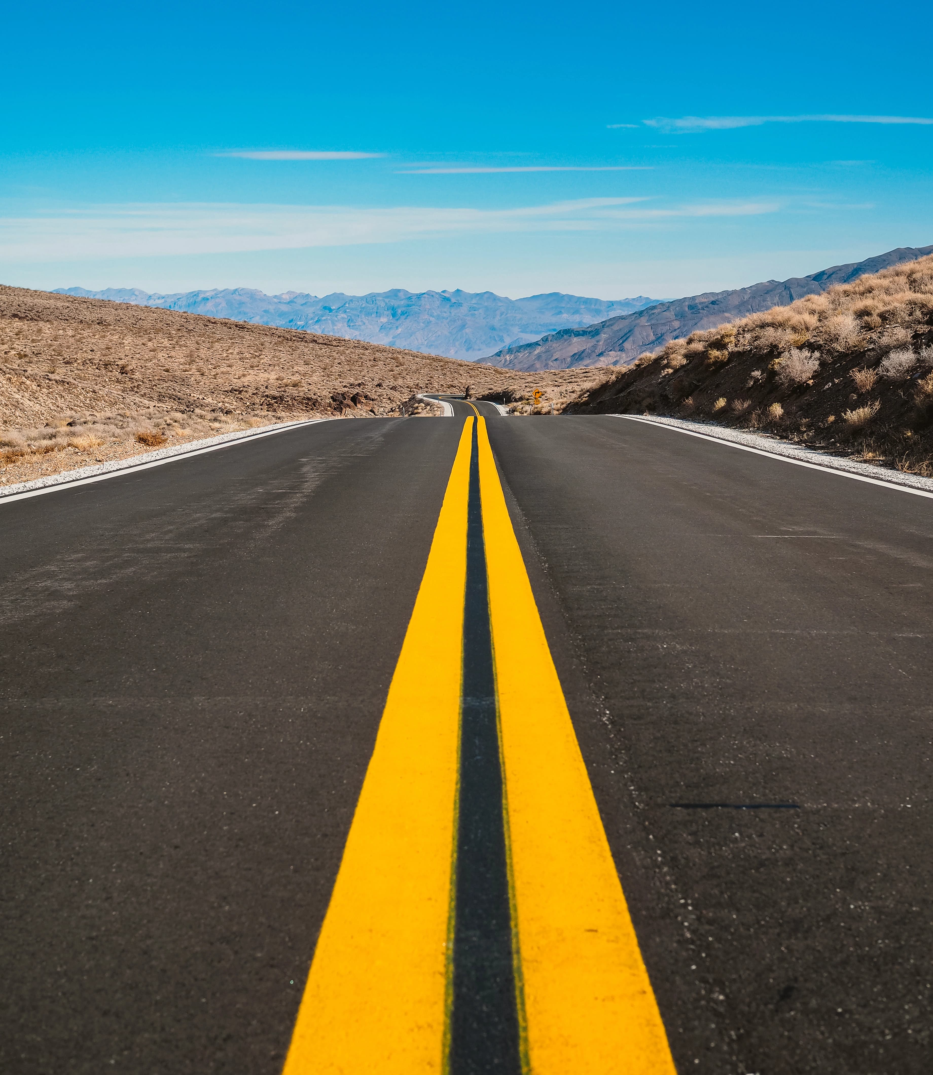 An empty desert highway photographed straight down its double yellow centre line towards the horizon, with the white WINCH W monogram floating over the asphalt.