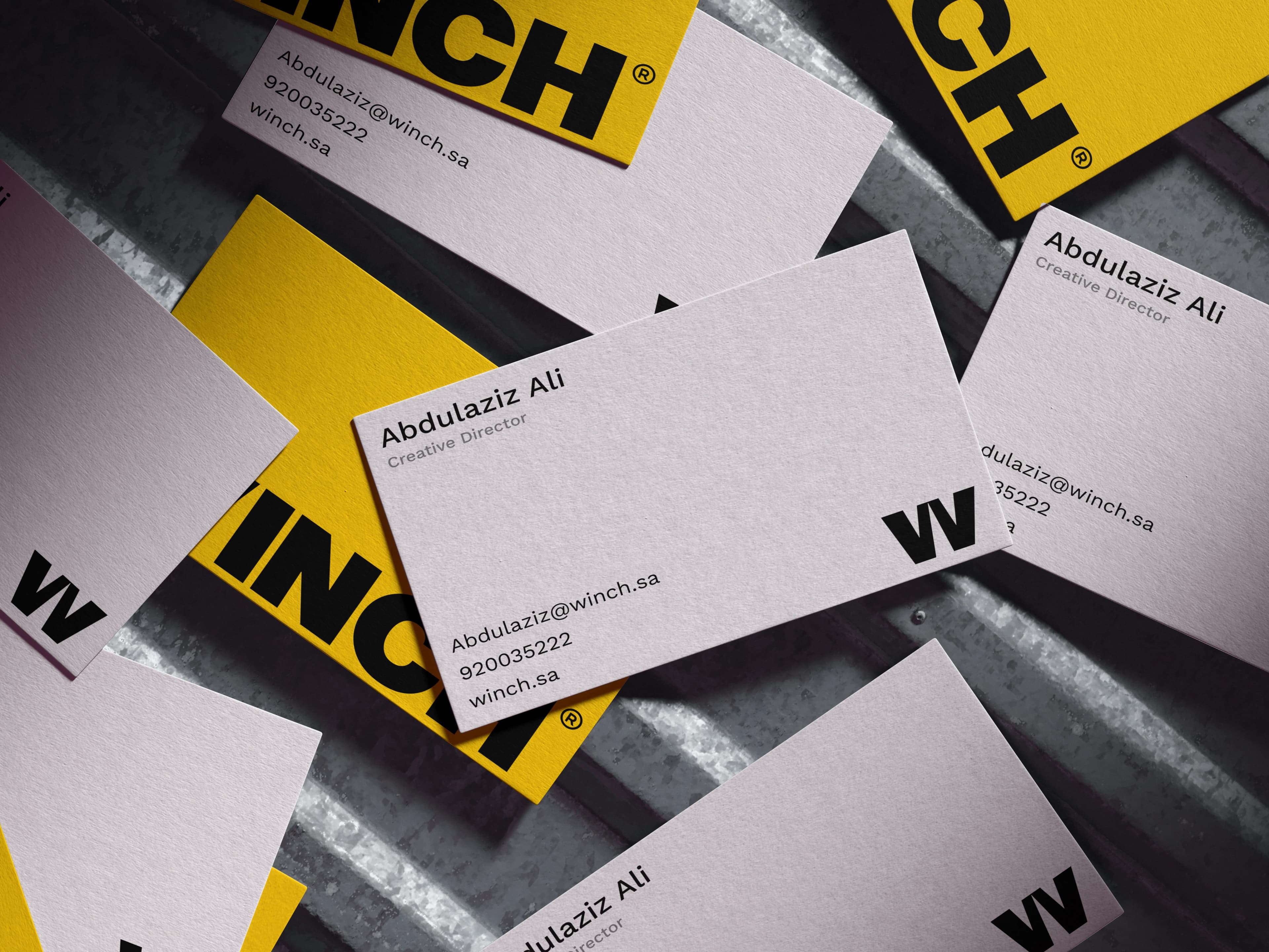 White and yellow WINCH business cards scattered across a brushed metal surface, the card in focus showing the W monogram above a name, job title, email address and phone number.