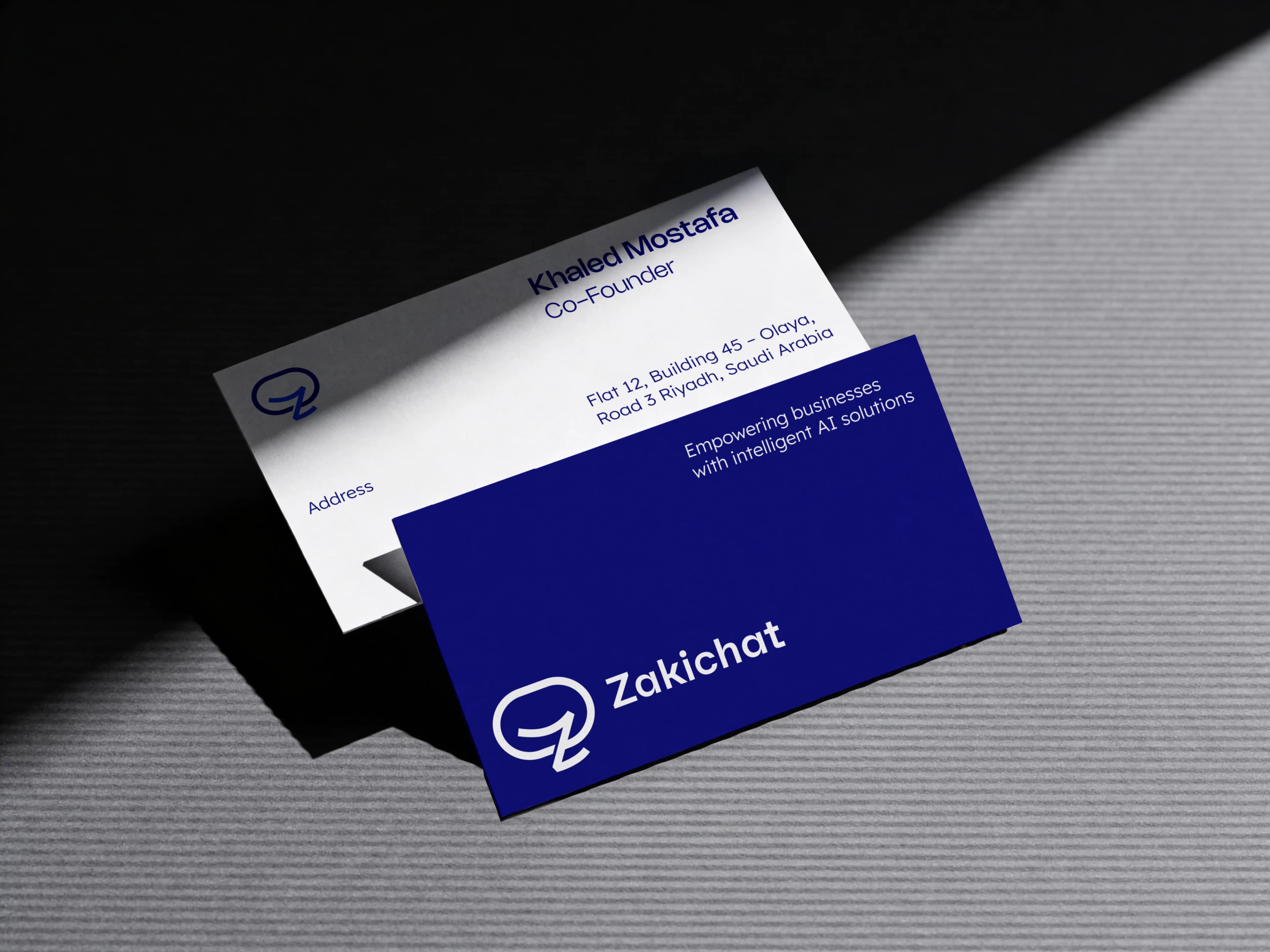 Zakichat — business cards A white and a navy Zakichat business card lying apart at different angles and partly overlapping, the white card carrying the name and a Riyadh address and the navy card the lockup with the line Empowering businesses with intelligent AI solutions.