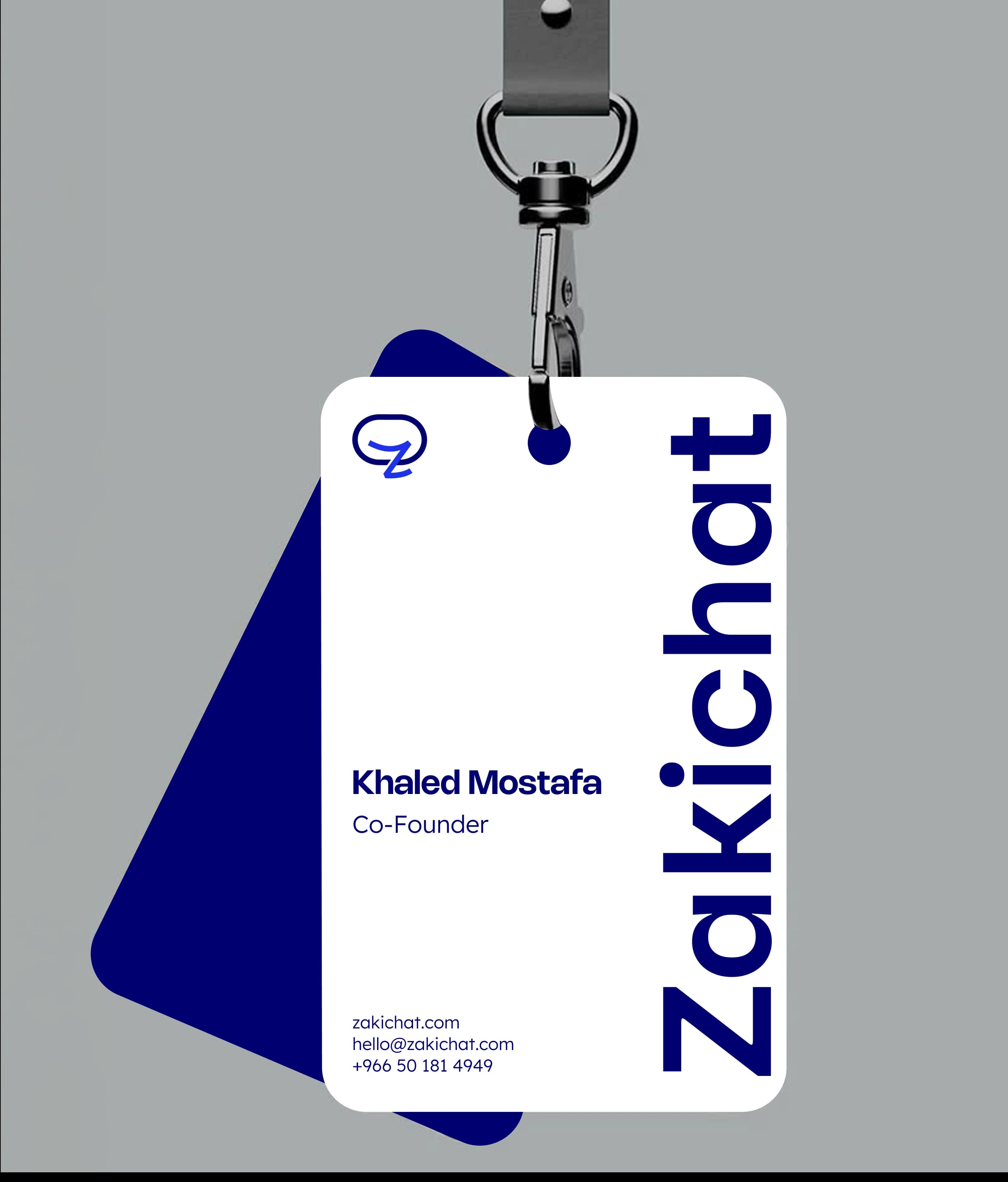 Zakichat — lanyard ID badge A white Zakichat ID badge on a metal clip over a flat grey-blue ground, printed with the mark, a large vertical Zakichat wordmark and the name Khaled Mostafa, Co-Founder, with a navy card angled behind it.