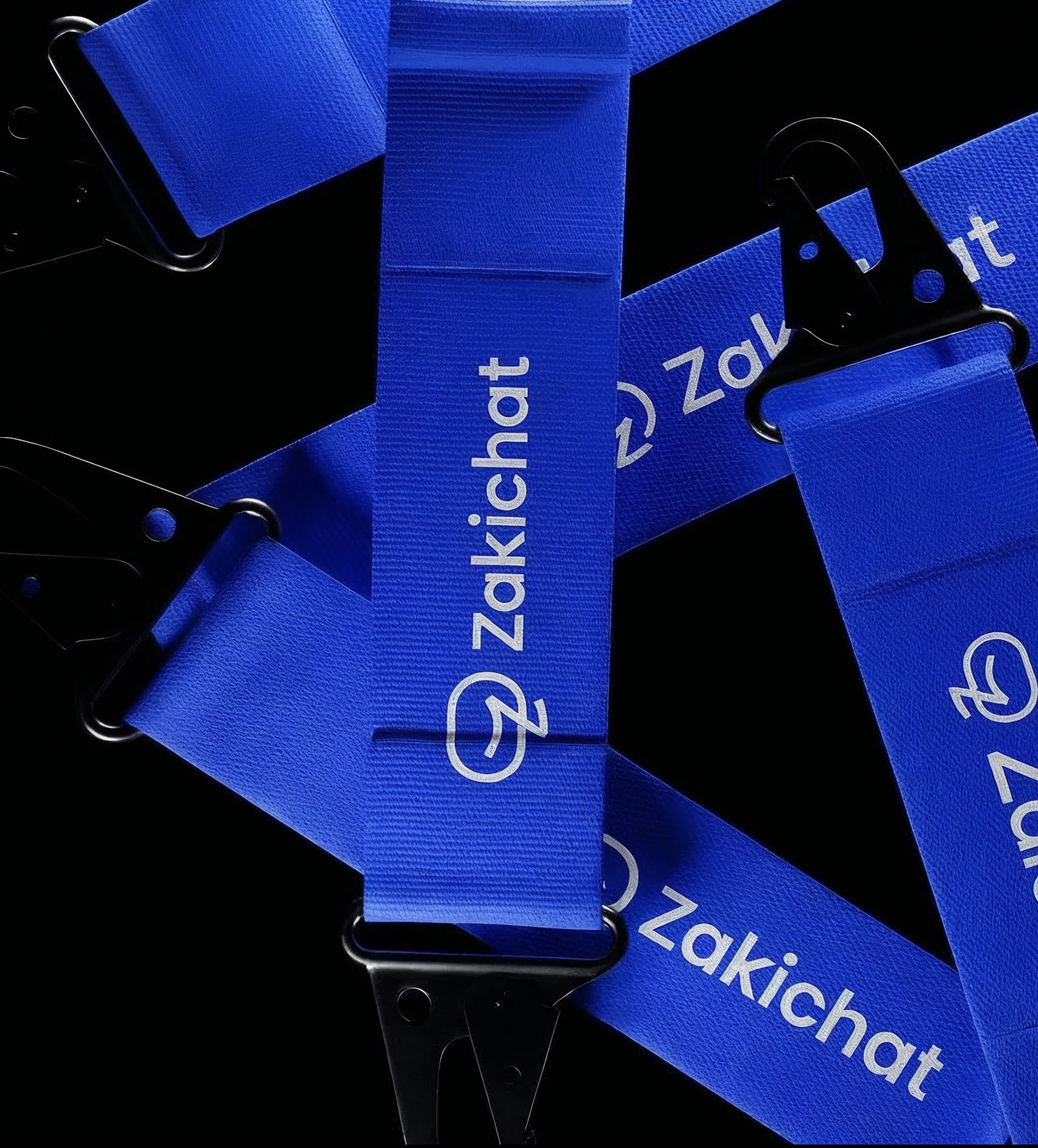 Zakichat — woven lanyards A pile of blue woven Zakichat lanyard straps crossing a near-black ground at angles, printed with the white mark and wordmark and fitted with black D-rings and swivel clips.