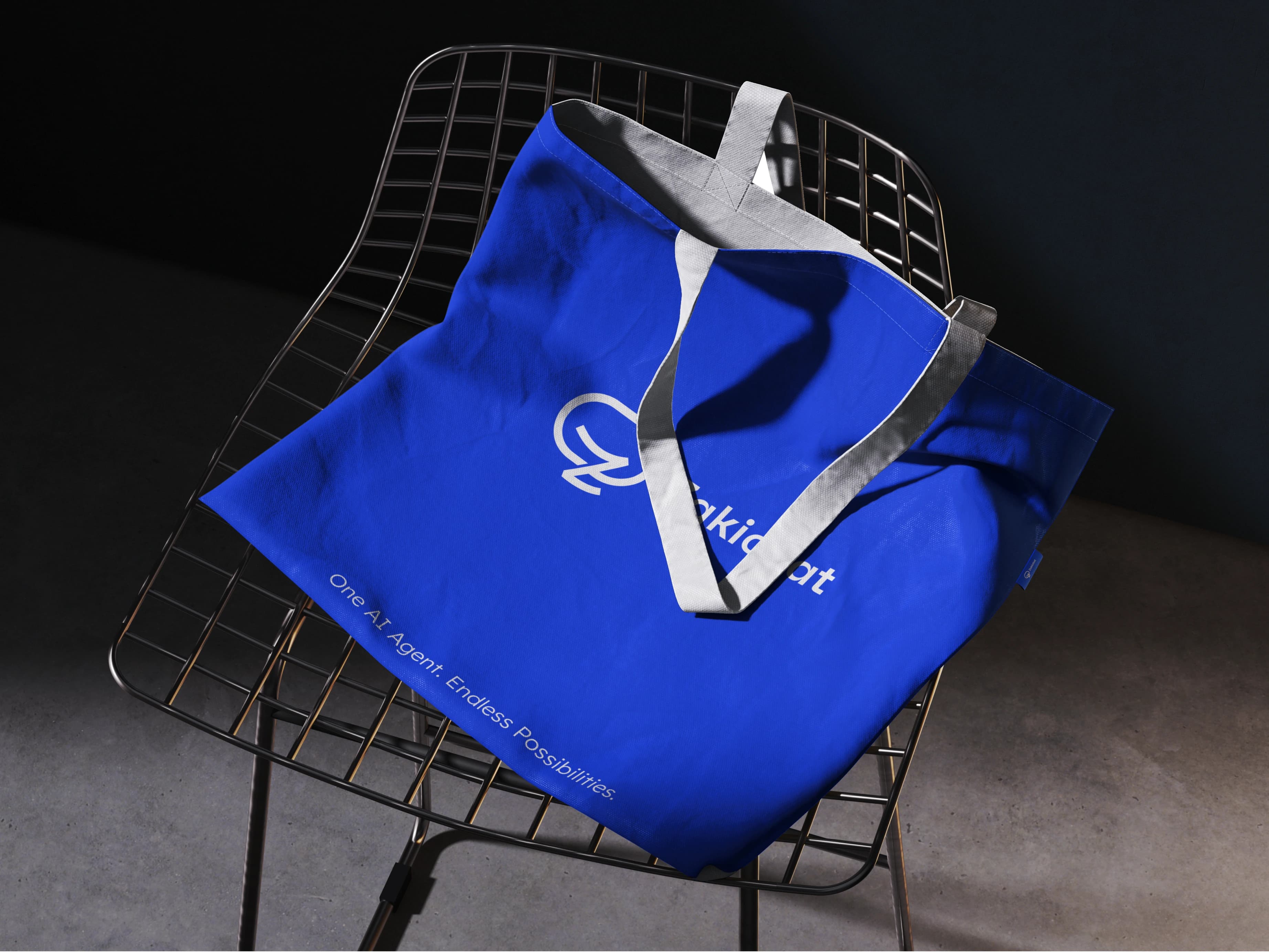 Zakichat — tote bag on chair A royal-blue Zakichat tote bag with grey webbing handles draped over a black wire-mesh chair in a dark studio, the handles throwing a hard shadow across the printed mark and wordmark.
